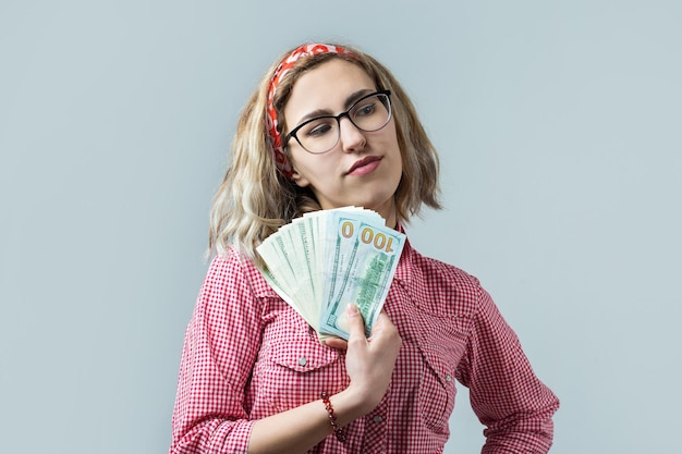 Gros plan d'une belle jeune femme dans une chemise à carreaux rouge dans des verres avec de l'argent en dollars américains dans la main sur fond gris