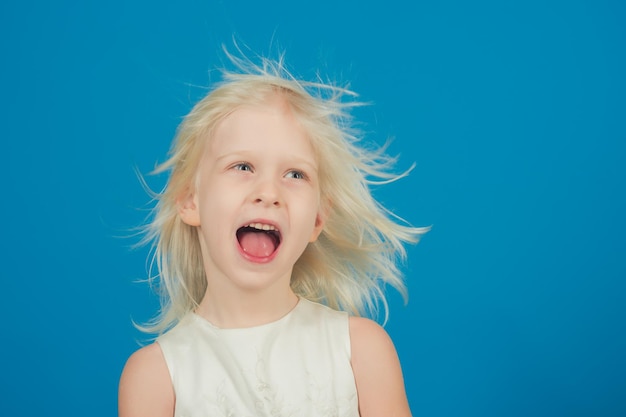 Gros plan d'une belle petite fille caucasienne blonde avec un sourire adorable posant contre le mur bleu de l'espace de copie Concept d'enfance heureuse