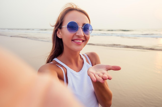 Gros plan d'une belle touriste qui profite du temps libre en plein air près de la plage de l'océan, regarde la caméra pendant les loisirs les jours d'été ensoleillés en faisant un selfie. Heureux touriste souriant dans un baiser d'air des tropiques