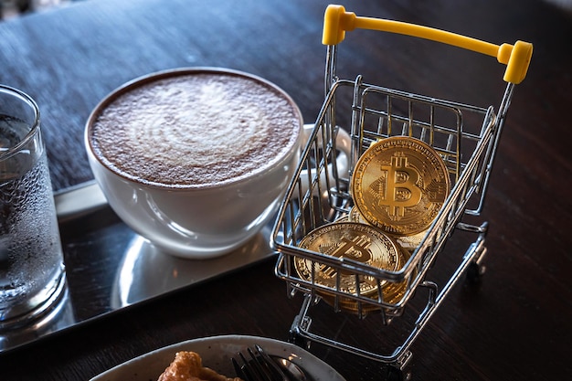 Gros plan sur des bitcoins dorés ou des pièces de crypto-monnaie ou des symboles dans le panier et du café au lait chaud avec de la mousse de lait latte art en forme de cygne dans une tasse de tasse sur la table Concept de monnaie future