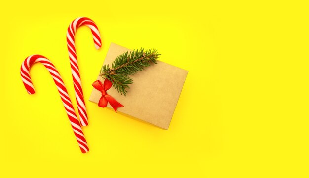 Gros plan d'une boîte-cadeau artisanale avec arc rouge, branche de sapin et bonbons, se trouve sur fond jaune avec espace de copie.