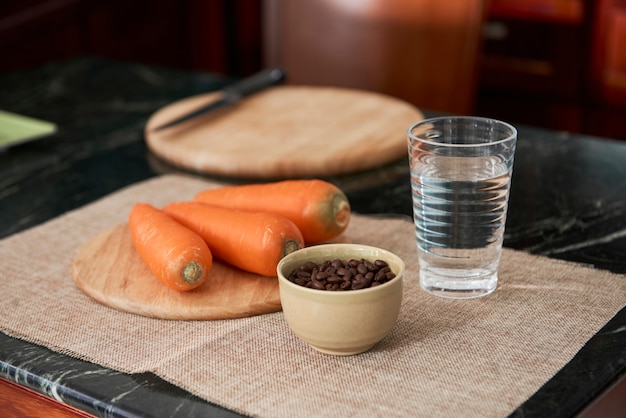 Gros plan sur un bol avec des grains de café, des carottes fraîches propres sur une planche à découper de forme ronde, un verre d'eau sur une serviette placée sur la table de la cuisine