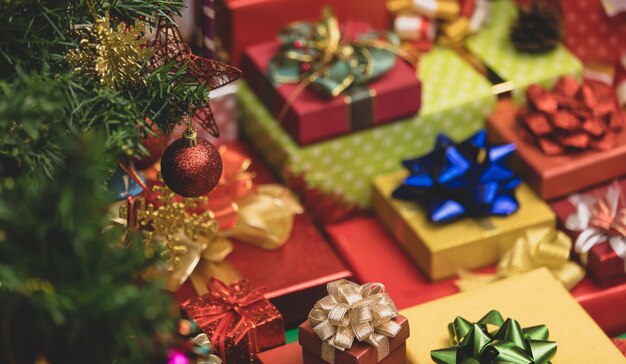 Gros plan d'une boule de sphère rouge brillante suspendue à la décoration sur une branche de pin de Noël devant un lot de boîtes-cadeaux enveloppées pour la veille de Noël empilées de rubans colorés et d'arcs en arrière-plan flou.
