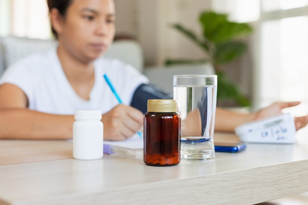 Gros plan sur une bouteille de pilules et un verre d'eau pendant qu'une jeune femme asiatique vérifie elle-même la pression artérielle et la fréquence cardiaque avec un manomètre numérique à la maison. Concept de santé et médical.