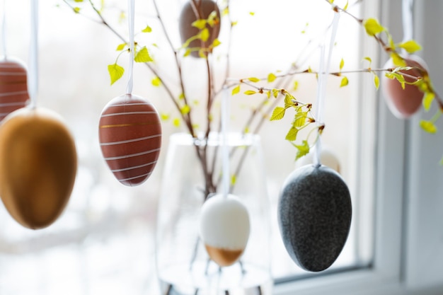 Gros plan des branches de printemps en fleurs de bouleau décorées d'oeufs de pâques dans un vase près de la fenêtre