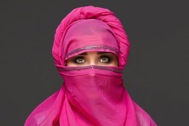 Gros plan d'une charmante femme au maquillage lumineux portant le hijab rose orné de paillettes. Elle pose au studio et regarde la caméra sur un fond sombre. Émotions humaines, faci