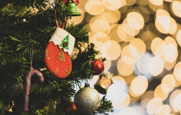 Gros plan d'une chaussette de décoration de réveillon de Noël rouge et blanche suspendue à la décoration sur une branche de pin de Noël vert avec une boule de sphère de bâton brillant et une cloche devant un arrière-plan flou de bokeh doré à faible luminosité.