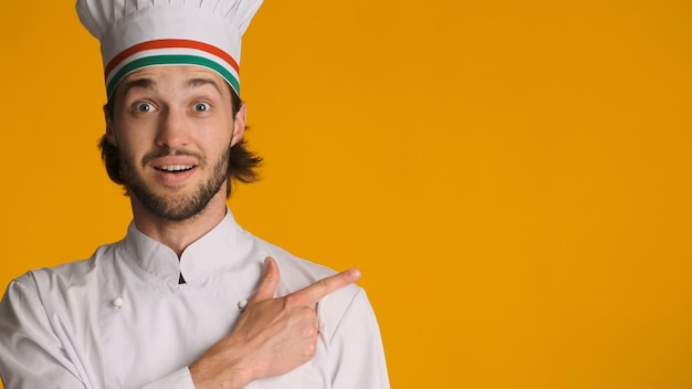 Gros plan chef surpris portant l'uniforme pointant vers l'espace pour la publicité ou la promotion sur fond coloré Homme étonné en toque montrant l'espace pour le texte