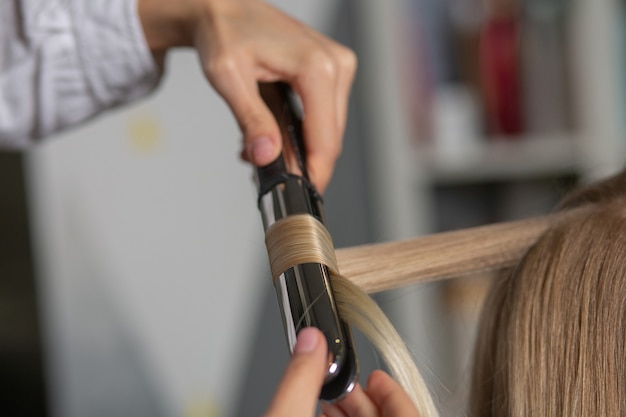 Gros plan d'un coiffeur faisant des boucles avec un fer à coiffer pour une femme blonde au salon de coiffure