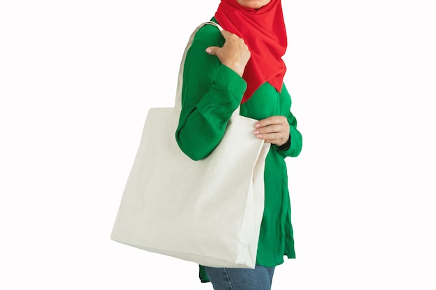 Gros plan d'une consommatrice musulmane tenant un sac fourre-tout écologique blanc vierge posant sur un espace de copie de fond de studio blanc