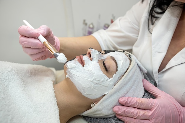 Gros plan d'une cosmétologue appliquant un masque facial cosmétique blanc pour sa patiente au salon de spa Concept de procédure cosmétique anti-rides