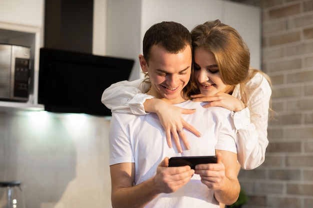 Gros plan d'un couple amoureux à l'aide d'un téléphone portable ensemble dans la cuisine. Femme aux cheveux longs, étreignant un homme par derrière