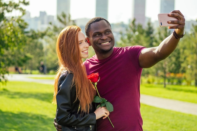 Gros plan d'un couple interracial tenant une rose blanche au printemps automne parc prenant un selfie avec un appareil photo de téléphone intelligent