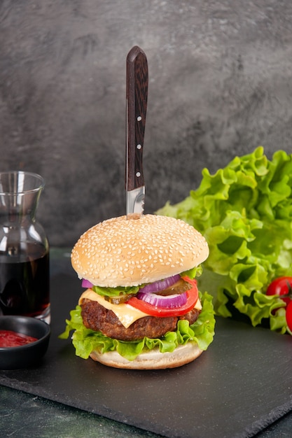 Gros plan d'un couteau dans un délicieux sandwich à la viande et du poivre vert sur un plateau noir sauce ketchup tomates avec tige sur une surface grise