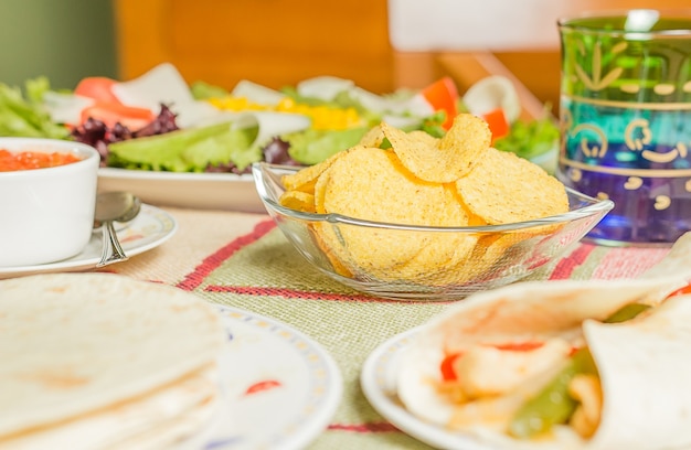 Gros plan sur une cuisine mexicaine traditionnelle dans une table, avec un bol de nachos, une sauce épicée, une assiette de fajita au poulet, des tortillas et une salade fraîche