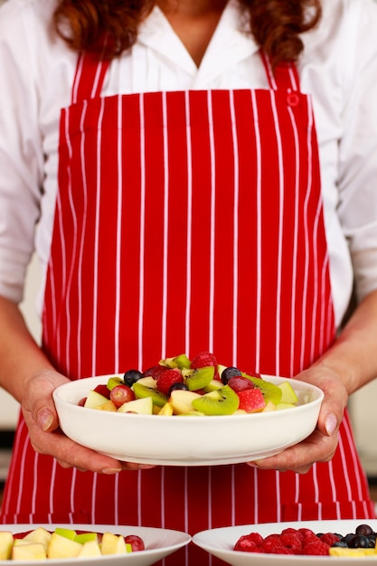 Gros Plan De Délicieux Délicieux Délicieux Apéritif De Salade De Fruits Mélangés Sucrés Pomme Kiwi Myrtille Raisin Dans Un Bol Blanc Sur Les Mains D'une Femme Chef Exécutive Qui Se Tient Debout Porte Un Tablier Rouge à Rayures Derrière En Arrière-plan.