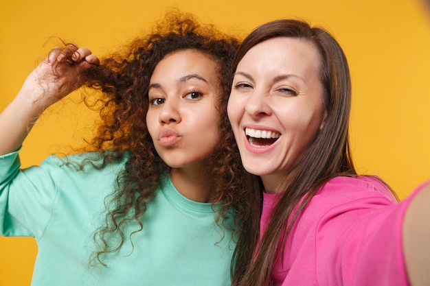 Gros plan de deux femmes amies fille afro-américaine européenne en vêtements verts roses posant isolés sur fond jaune. Concept de style de vie des gens. Maquette de l'espace de copie. Faire selfie tourné sur téléphone mobile.