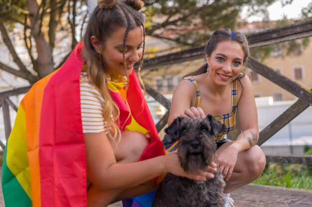 Gros plan sur deux jeunes femmes de race blanche tenant un drapeau de fierté LGBT dans un parc à côté de leur chien noir