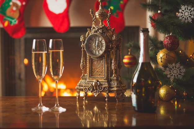 Gros plan de deux verres de champagne sur la table à côté de la vieille horloge montrant minuit au salon décoré pour Noël