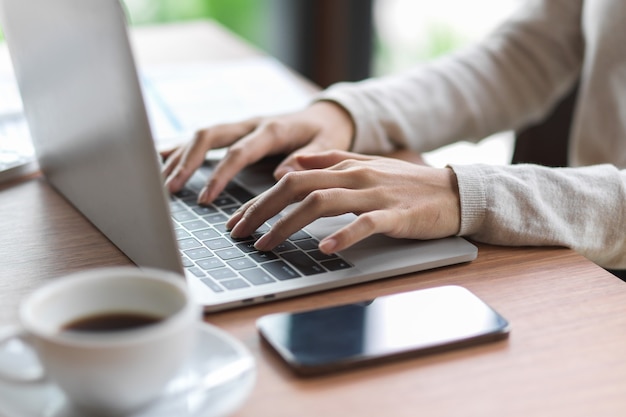 Gros plan sur des doigts féminins tapant sur un clavier d'ordinateur portable avec un smartphone et du café sur le bureau en bois