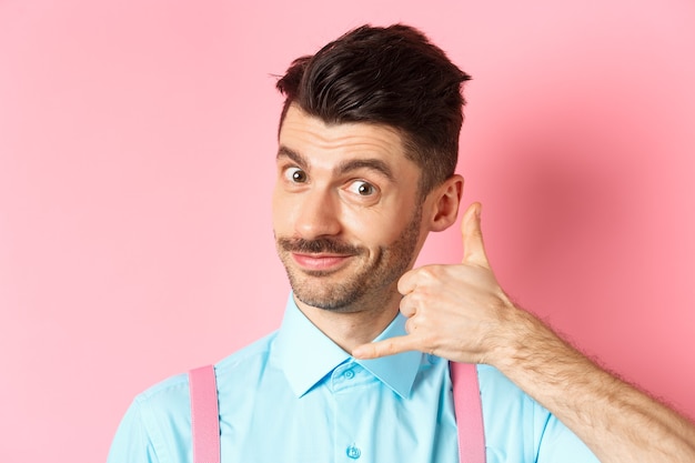 Gros plan du beau jeune homme avec moustache, montrant le geste du téléphone, demandant à l'appeler, debout sur le rose.