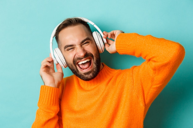 Gros plan du bel homme moderne, écouter de la musique dans les écouteurs, debout en pull orange sur fond turquoise.