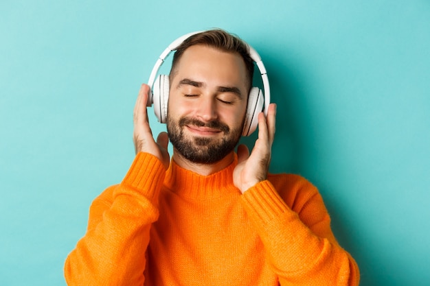 Gros plan du bel homme moderne, écouter de la musique dans les écouteurs, debout en pull orange sur fond turquoise.