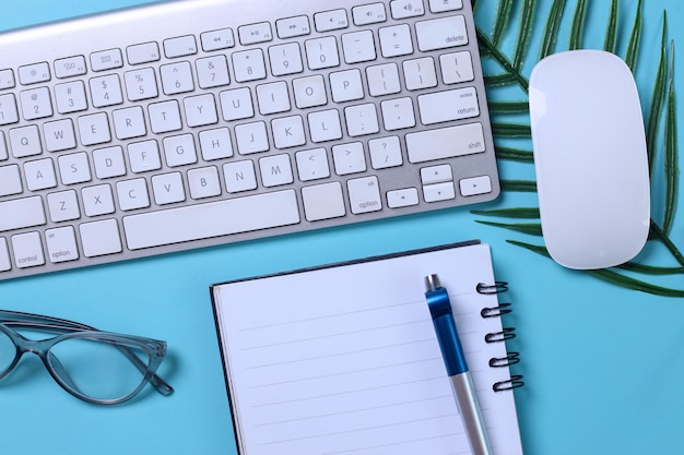 Gros plan du bureau avec clavier, lunettes de souris en feuille de palmier et ordinateur portable sur fond bleu