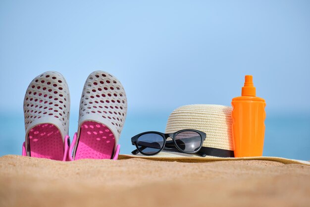 Gros plan du chapeau de paille sabots chaussures crème solaire et lunettes de soleil de protection noires sur la plage de sable au bord de la mer tropicale par une chaude journée ensoleillée concept de vacances d'été