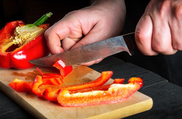 Gros plan du chef ou cuisinier mains coupant des poivrons sur une planche à découper. Préparation professionnelle de salade dans la cuisine d'un restaurant ou d'un café. Vue de côté