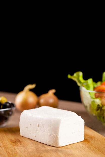 Gros plan sur du fromage feta allongé sur une planche en bois à côté d'oignons, de salade fraîche et d'un bol en verre d'olives sur fond noir