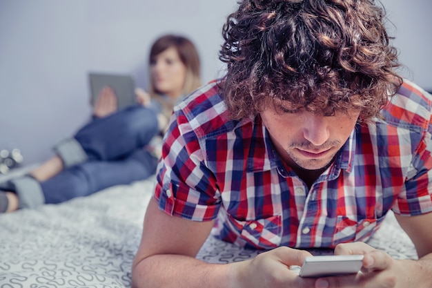 Gros plan du jeune bel homme utilisant un smartphone allongé sur le lit et belle femme lisant dans une tablette électronique. Concept de temps libre à la maison.