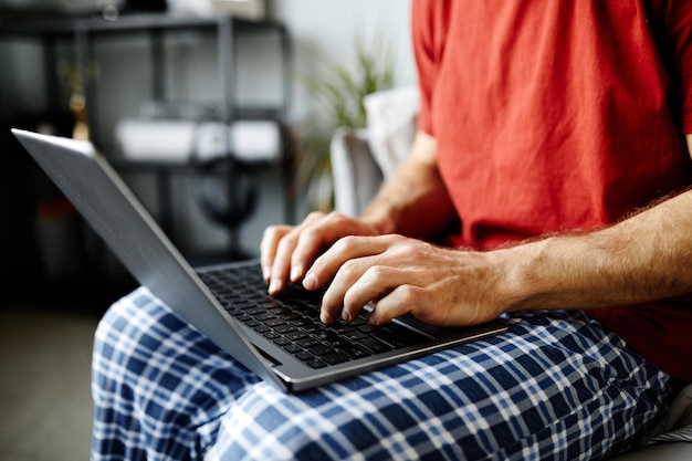 Gros plan du jeune homme assis sur le lit avec un ordinateur portable sur ses genoux et en tapant dessus