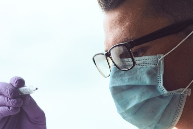 Gros plan du jeune homme portant des lunettes, un masque médical et des gants jetables en regardant thermomètre.