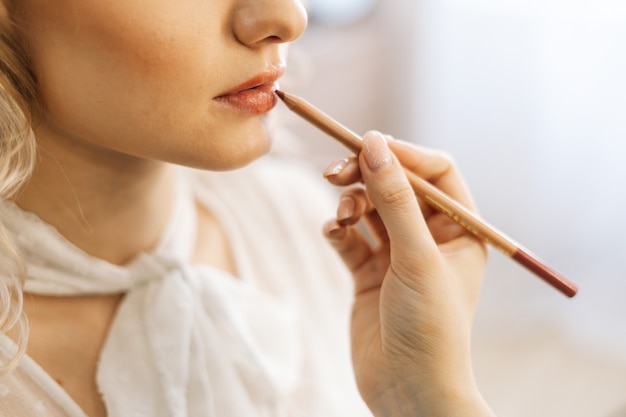 Gros plan du maquilleur professionnel faisant le maquillage des lèvres de la jeune femme avec un crayon à lèvres