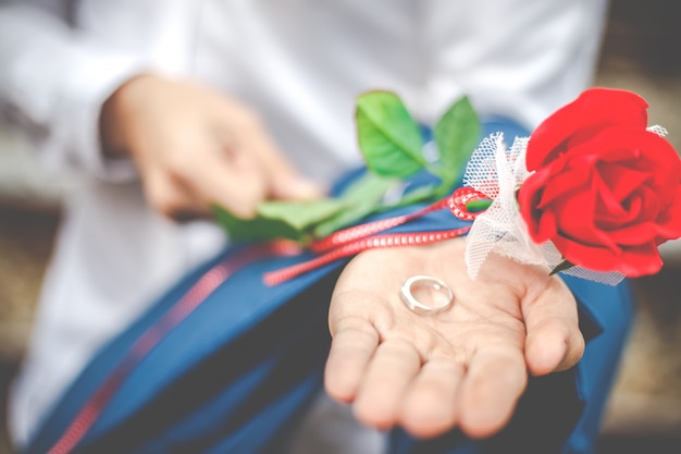 Gros plan du marié tenant la bague de mariage sur la paume avec rose rouge