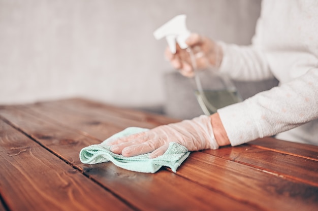 Gros plan du nettoyage de la table en bois à la maison, de la désinfection de la surface de la table de la cuisine avec un flacon pulvérisateur désinfectant antibactérien, du lavage des surfaces avec une serviette et des gants.