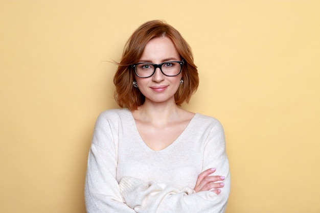 Gros plan du portrait en studio de joyeuse femme réussie satisfaite avec les cheveux courts porte des lunettes, pull surdimensionné blanc, poses, souriant, bras croisés, regardant la caméra. Isolé sur une surface beige