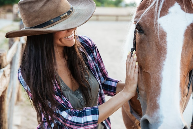 Gros plan du sourire tendre jeune femme cow-girl au chapeau avec son cheval