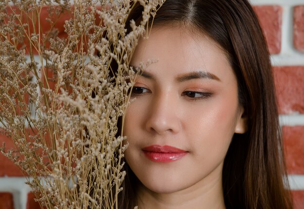 Gros plan du visage du jeune beau modèle féminin asiatique attrayant avec la branche de la fleur sur fond de mur de brique orange.