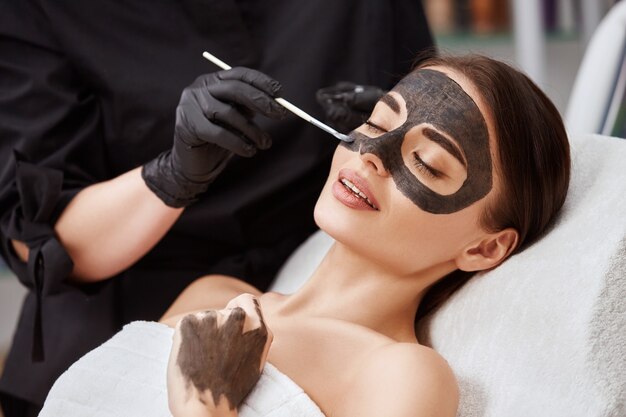 Gros plan du visage de femme couchée dans le salon et esthéticienne appliquant un masque de carbone, belle femme ayant des procédures de soins de la peau dans le spa