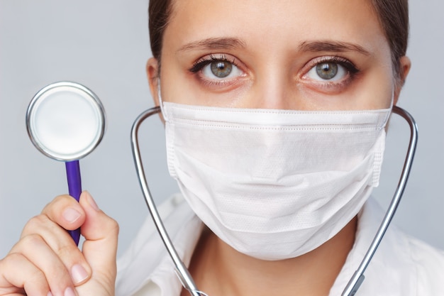 Gros plan du visage d'une femme médecin avec un stéthoscope dans un masque médical sur fond gris