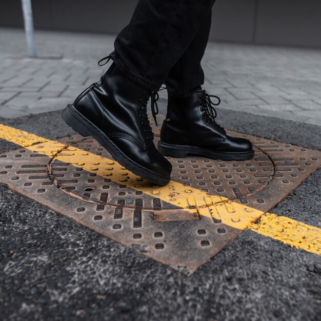 Gros plan d'élégantes bottes automne-printemps en cuir sur les jambes des hommes. Jeune homme en jeans vintage dans des chaussures à la mode se dresse sur un trou d'homme vintage en fer dans la ville. Mode jeunesse. Chaussures à la mode pour hommes.