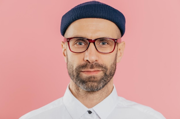 Gros plan d'un employé masculin confiant avec une barbe épaisse et une moustache porte des lunettes transparentes chapeau chemise blanche se dresse sur fond rose a un regard attentif sur la caméra Expressions faciales