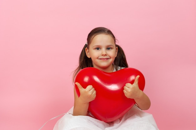 Gros plan enfant souriant en robe blanche tenant un ballon rouge en forme de coeur, donnant les pouces vers le haut