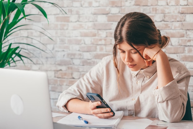 Gros plan sur une étudiante du millénaire assise au bureau, tenant le téléphone à la main, vérifiant des informations ou des données importantes en prenant des notes sur papier Jeune fille concentrée se préparant à un test ou à un examen universitairexA