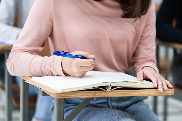 Gros plan sur les étudiants universitaires écrivant une note sur un cahier dans la salle de classe tout en écoutant et en étudiant la conférence. Gros plan des mains de la femme écrivant sur le bloc-notes placé sur un bureau en bois