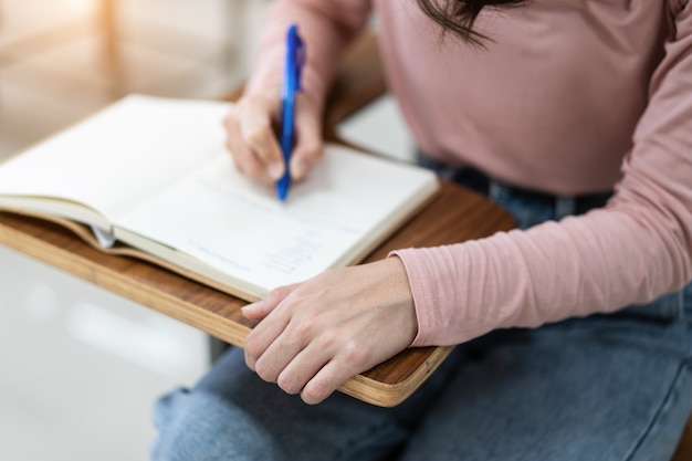 Gros plan sur les étudiants universitaires écrivant une note sur un cahier dans la salle de classe tout en écoutant et en étudiant la conférence. Gros plan des mains de la femme écrivant sur le bloc-notes placé sur un bureau en bois