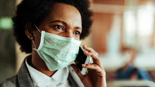 Gros plan d'une femme d'affaires afro-américaine portant un masque facial pour la protection contre les virus tout en parlant au téléphone à l'aéroport