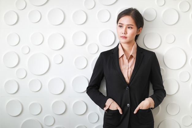 Gros plan d'une femme d'affaires asiatique debout, souriante et glissant la main dans le sac du costume qui montre le confort d'être photographié sur un fond comportant beaucoup de surface circulaire blanche.